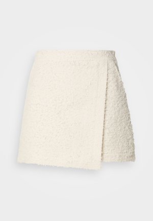 Mini-jupe en tissu beige crème texturé avec un devant croisé asymétrique et une ceinture droite sur un fond uni.
