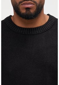 Pull en maille noire avec un col ras du cou côtelé et un tissu texturé, présentant une vue rapprochée de l'encolure et de la partie supérieure de la poitrine.