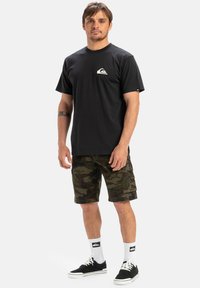 Black cotton t-shirt with a white logo on the left chest, paired with camouflage cargo shorts, white socks, and black sneakers.