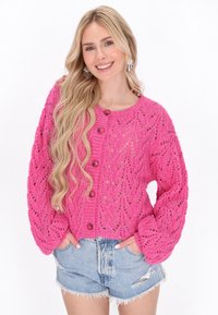 Rožinė megzta kardiganas su tekstūriniu raštu, apvaliu iškirpimu, ilgomis rankovėmis ir keturiais mygtukais, derinamas su džinsiniais šortais.