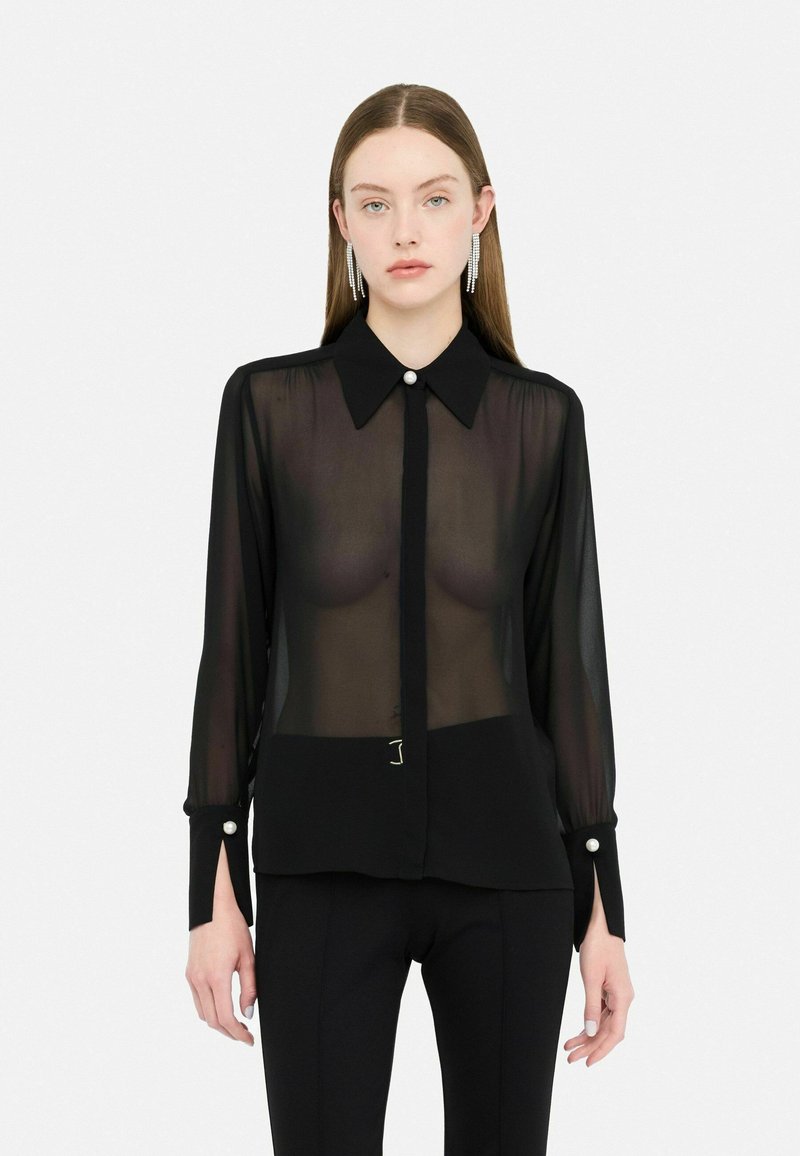 Blusa nera trasparente con maniche lunghe, colletto classico, dettagli con bottoni perla e spacchi laterali; abbinata a pantaloni neri a vita alta.