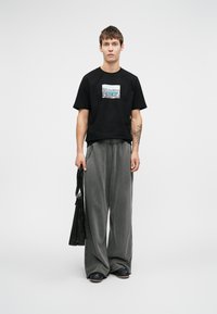 Tânăr îmbrăcat cu un tricou grafic negru, pantaloni gri largi, pantofi negri, ținând o geantă neagră, stând în fața unui fundal alb simplu.