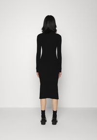 Abito a maglia nero a coste con maniche lunghe, silhouette aderente e lunghezza midi, abbinato a scarpe nere e calze su uno sfondo neutro.
