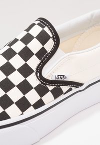 Zwart-wit geblokte slip-on sneaker met canvas bovenwerk, gevoerde kraag, elastische zijaccenten en rubberen zool. "VANS" label aan de zijkant.