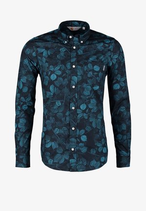 Mand gående iført en sort blazer, blomstret skjorte med knapper, mørke jeans med opbuk, grå sko, bærende på en grøn og brun lædertaske.