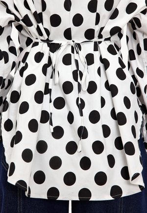 Blouse en tissu blanc ornée de grands pois noirs, avec une taille froncée maintenue par un mince cordon blanc et des manches amples.