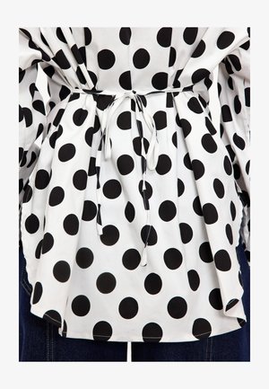 Blouse en tissu blanc ornée de grands pois noirs, avec une taille froncée maintenue par un mince cordon blanc et des manches amples.