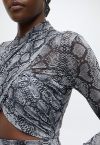 Haut à manches longues avec imprimé serpent gris et noir, présentant des fronces sur le côté et un col en style cache-cœur. Confectionné dans un matériau transparent.