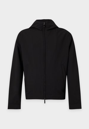 Homme portant une veste noire zippée, une chemise blanche texturée, un jean bleu déchiré et des baskets blanches, debout avec une main dans la poche.