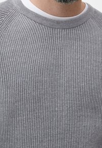 Mann in grauem geripptem Pullover über einem weißen Rundhalsausschnitt-Shirt, das seinen Hals und einen Teil seines grauen Bartes zeigt.