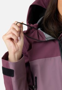 Vandtæt jakke i mørk maroon og lys lilla, med sort justerbart snor, lynlåslukning og Velcro-manchet detaljer.