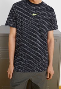 Černé tričko s opakujícím se šedým vzorem loga Nike swoosh, kulatým výstřihem, krátkými rukávy a neonově zeleným logem Nike na hrudi.