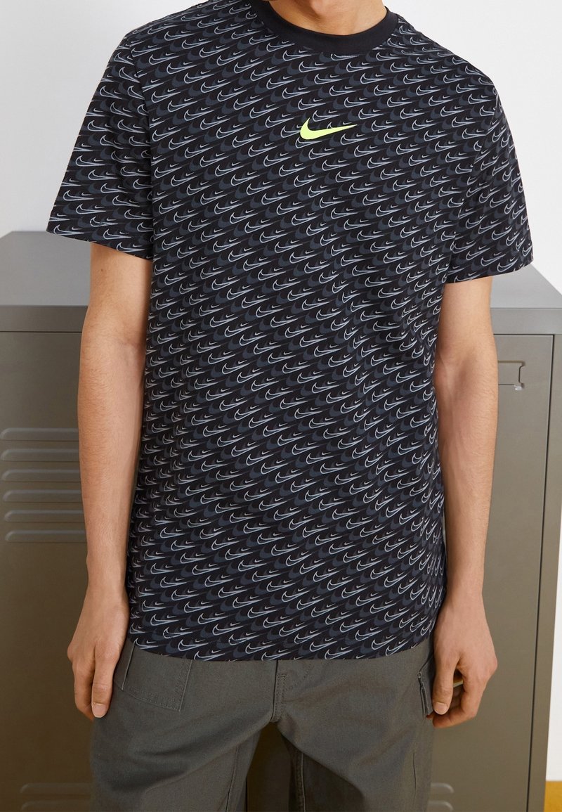 Černé tričko s opakujícím se šedým vzorem loga Nike swoosh, kulatým výstřihem, krátkými rukávy a neonově zeleným logem Nike na hrudi.
