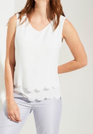 Kobieta ubrana w białą bluzkę bez rękawów z falowanym dołem oraz jasne spodnie, stojąca z jedną ręką na biodrze na tle jednolitego tła.