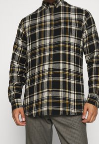 Camisa de cuadros en blanco y negro con acentos amarillos, con cuello abotonado, mangas largas y botones marrones. Textura suave de franela.