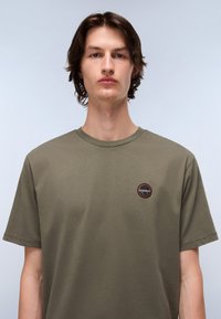 Tricou scurt cu mânecă scurtă de culoare verde măsliniu, realizat din bumbac, având un patch cu logo rotund pe pieptul stâng, cu un decolteu clasic și croială lejeră.
