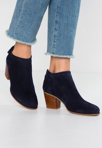 Stivaletti da donna in camoscio blu navy con punta rotonda, tacco basso in legno impilato e chiusura con cerniera sul retro. Design aderente con linee pulite.