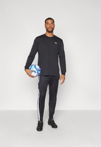 Una camiseta negra de manga larga y pantalones a juego con rayas blancas a los lados. El modelo sostiene un balón de fútbol multicolor, con zapatos negros.