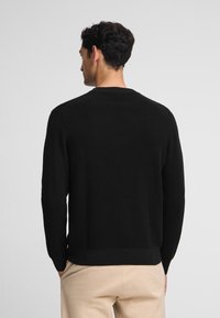 Suéter negro de punto con escote redondo, que presenta un patrón texturizado y puños acanalados. Combinado con pantalones de color claro.