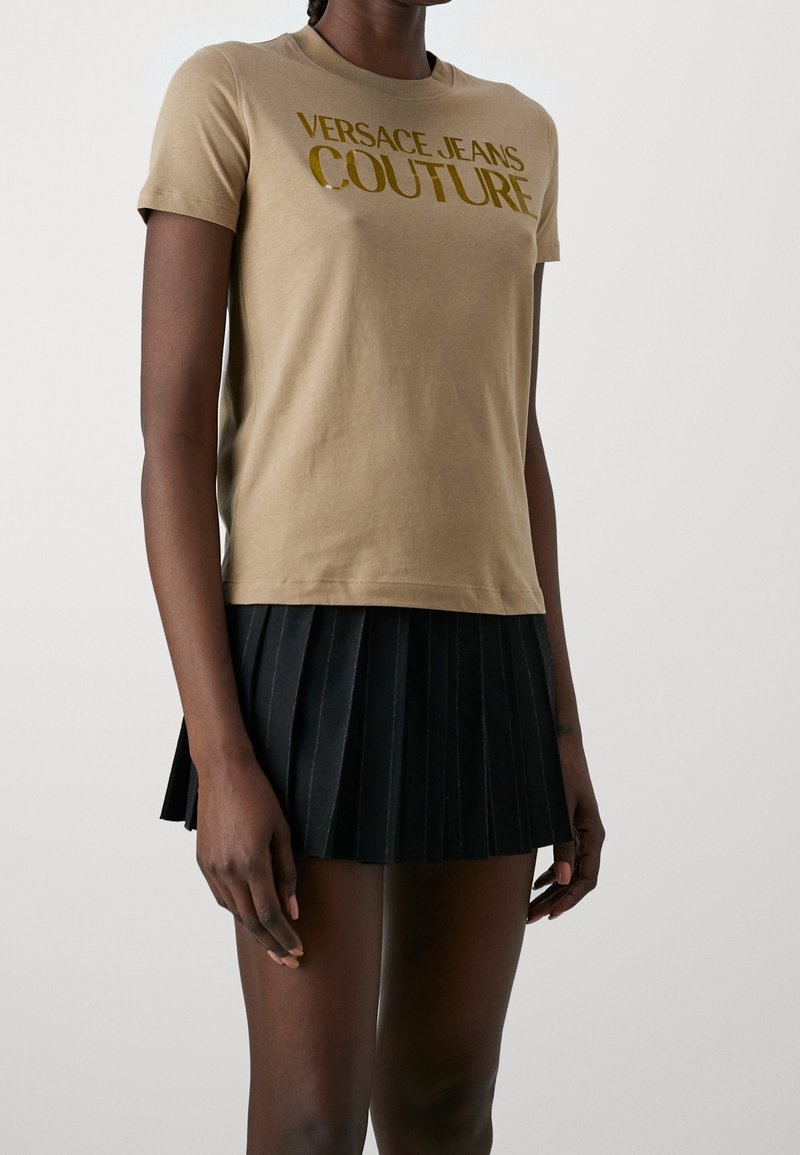 Beige katoenen t-shirt met korte mouwen met gouden reflecterende tekst "Versace Jeans Couture". Gecombineerd met een zwarte geplooide rok.