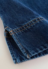 Gros plan sur l'ourlet d'un jean en denim bleu avec des coutures orange détaillées et une texture de tissu légèrement usée sur un fond clair.