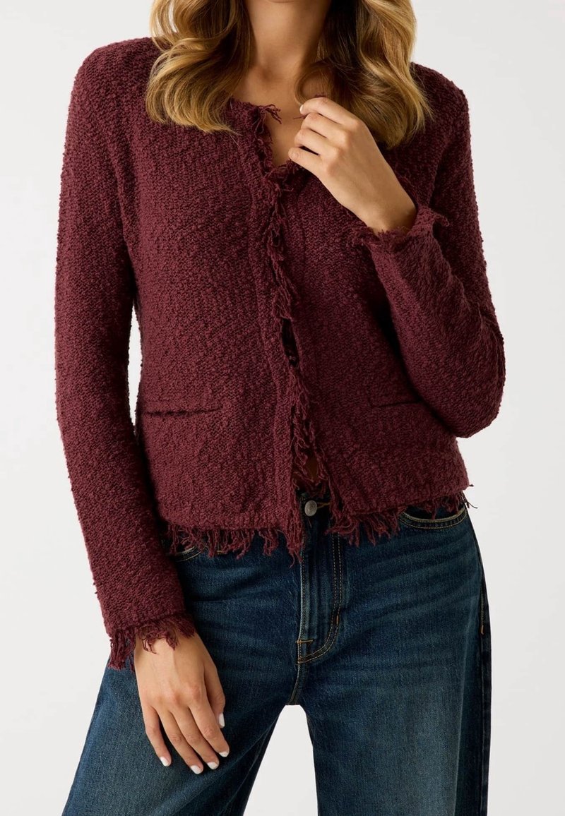 Femme portant un cardigan bordeaux texturé aux bords effilochés, sur un jean bleu foncé, visible du cou aux hanches, sur un fond neutre.