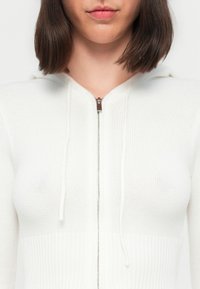 Frau mit schulterlangem braunem Haar, die einen weißen, gestrickten Zip-Hoodie mit sichtbaren Kordeln in der Nähe der Schlüsselbeine trägt.