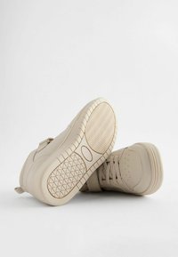 Baskets beige pour tout-petits avec lacets et une sangle velcro, montrant des semelles en caoutchouc texturé et une zone de orteil perforée sur un fond blanc.