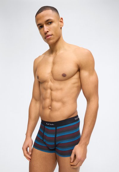 Μυώδης άνδρας με κοντά μαλλιά που φοράει μαύρα, μπλε και κόκκινα ριγωτά εσώρουχα τύπου μποξέρ, στέκεται μπροστά σε απλό φωτεινό φόντο.