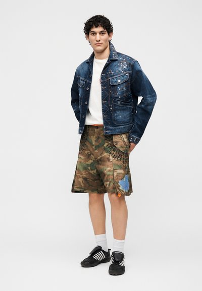 Νέος άνδρας που φοράει τζιν μπουφάν με καρφιά, λευκό μπλουζάκι, καμουφλάζ cargo σορτς με μπαλώματα, λευκές κάλτσες και μαύρα αθλητικά, στέκεται μπροστά σε απλό φόντο.