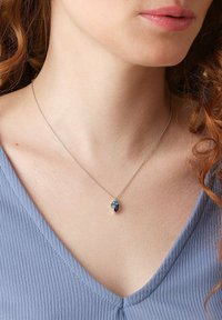 Silberne Kettenhalskette mit einem blauen Edelsteinanhänger im floralen Design, vor einem strukturierten hellblauen Oberteil.