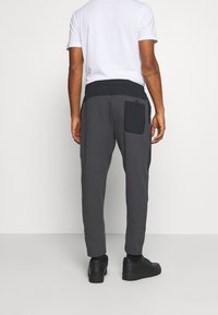 Man in donkergrijze joggers met een zwarte achterzak en tailleband, een wit t-shirt en zwarte sneakers, staande tegen een effen achtergrond.