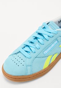 Světle modré semišové tenisky s perforovanou špičkou, kontrastními neonově žlutými akcenty a silnou gumovou podrážkou. Obsahují ploché tkaničky a logo.