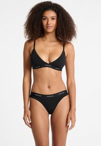 Black cotton bralette and matching bikini featuring a V-neck design, thin adjustable straps, and branding along the elastic waistband.