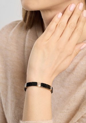 Bracelet noir avec des maillons carrés et des accents dorés, porté au poignet, sur un fond neutre.