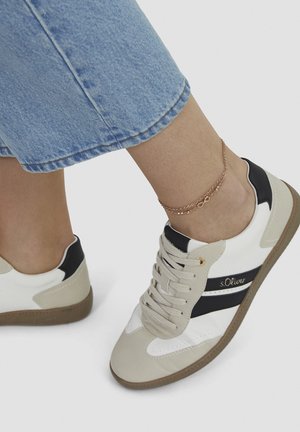 Beine in hellblauen Jeans, die beige und weiße Sneakers mit schwarzen Streifen und ein goldenes Fußkettchen auf weißem Hintergrund tragen.