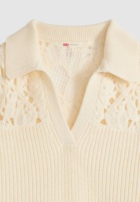 Pull en maille crème avec une texture côtelée, comportant une insertion en dentelle et un col en V. Design à col avec manches courtes. Étiquette taille S visible.