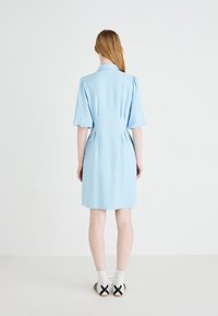 Robe bleu clair avec des manches courtes bouffantes, un col et un dos plissé, fabriquée en tissu lisse. Portée avec des chaussettes blanches et des chaussures avec des accents noirs.