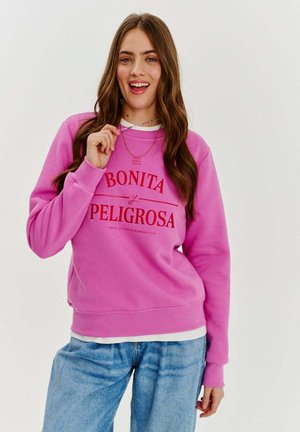 Różowy sweter z czerwonym napisem "BONITA y PELIGROSA." Posiada ściągacze przy mankietach i na dole, noszony na białej koszuli oraz dżinsowych spodniach.