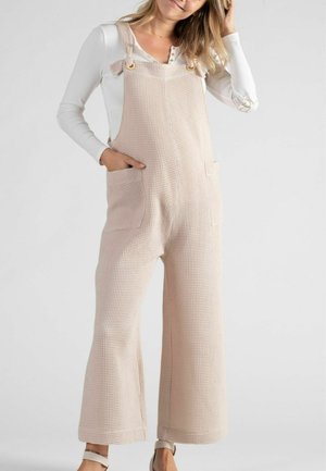 Salopette en rose pâle, texturée en gaufre, avec des jambes larges, des bretelles ajustables et des poches avant, portée avec une chemise à manches longues blanche.