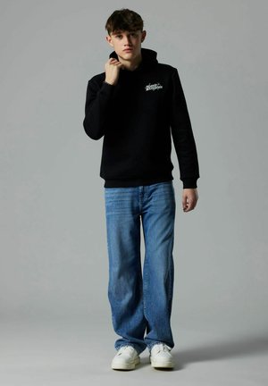 Giovane uomo che indossa un felpone nero con del testo, jeans blu e sneakers bianche, in piedi contro uno sfondo grigio uniforme.