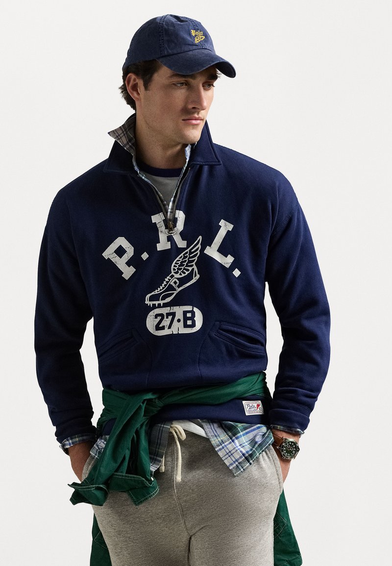 Pulover navy cu text și logo alb, guler cu fermoar, buzunare tip kangaroo, suprapus peste o cămașă în carouri; asociat cu pantaloni de trening gri și un pulover verde legat.