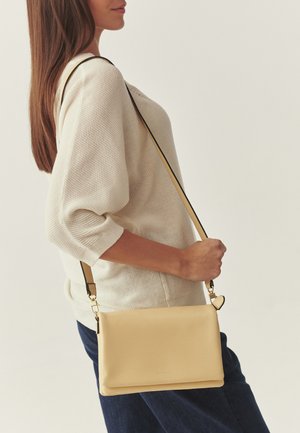 Frau trägt beigefarbenen Strickpullover und dunkle Hose, mit einer hellbraunen Leder-Umhängetasche mit schwarzem Riemen über der Schulter.