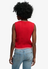 Pull sans manches rouge en tricot avec un motif torsadé texturé, bordé d'une bande rayée blanche à l'ourlet. Porté avec un jean bleu clair.