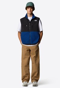 Czarny i niebieski polarowy bezrękawnik z zamykanymi kieszeniami, w połączeniu z khaki spodniami i szarymi trampkami. Model noszący zieloną dzianinową czapkę.