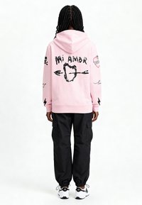 Felpa con cappuccio rosa con disegni grafici neri, tra cui "Mi Amor", cuori e motivi di teschi, abbinata a pantaloni cargo neri e sneakers bianche.