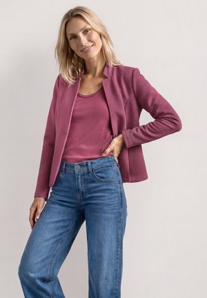 Smilende kvinde står med hånden på hoften iført en tekstureret rosa jakke, matchende top og blå jeans mod en neutral baggrund.