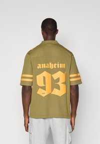 Olivově zelená košile s krátkým rukávem, na které je žlutý nápis "anaheim" a velké číslo "93" na zádech. Na rukávech jsou horizontální žluté pruhy.