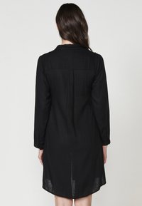 Rochie din in negru cu mâneci lungi și mansete cu nasturi, având un design cu umeri și pliu la spate, cu lungime până la genunchi și o siluetă dreaptă.