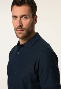 Navyblå pikétröja med ett texturerat randmönster, utrustad med krage, två knappar och en logotyp nära nederkanten. Mjuk tygkänsla.